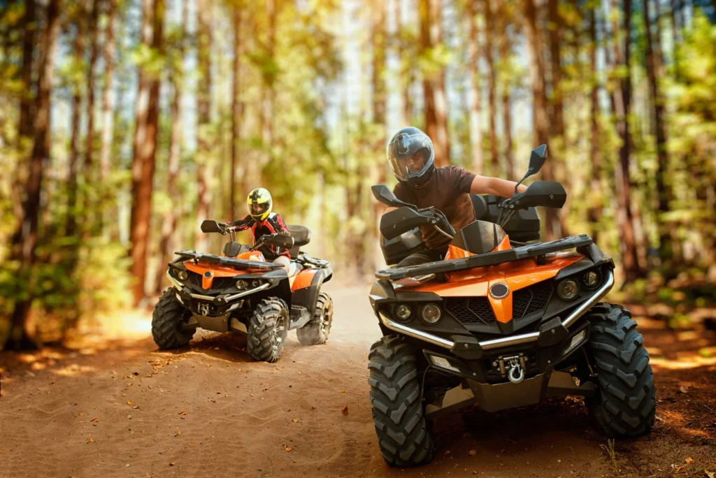 Two helmeted riders driving all-terrain vehicles on a wooded trail